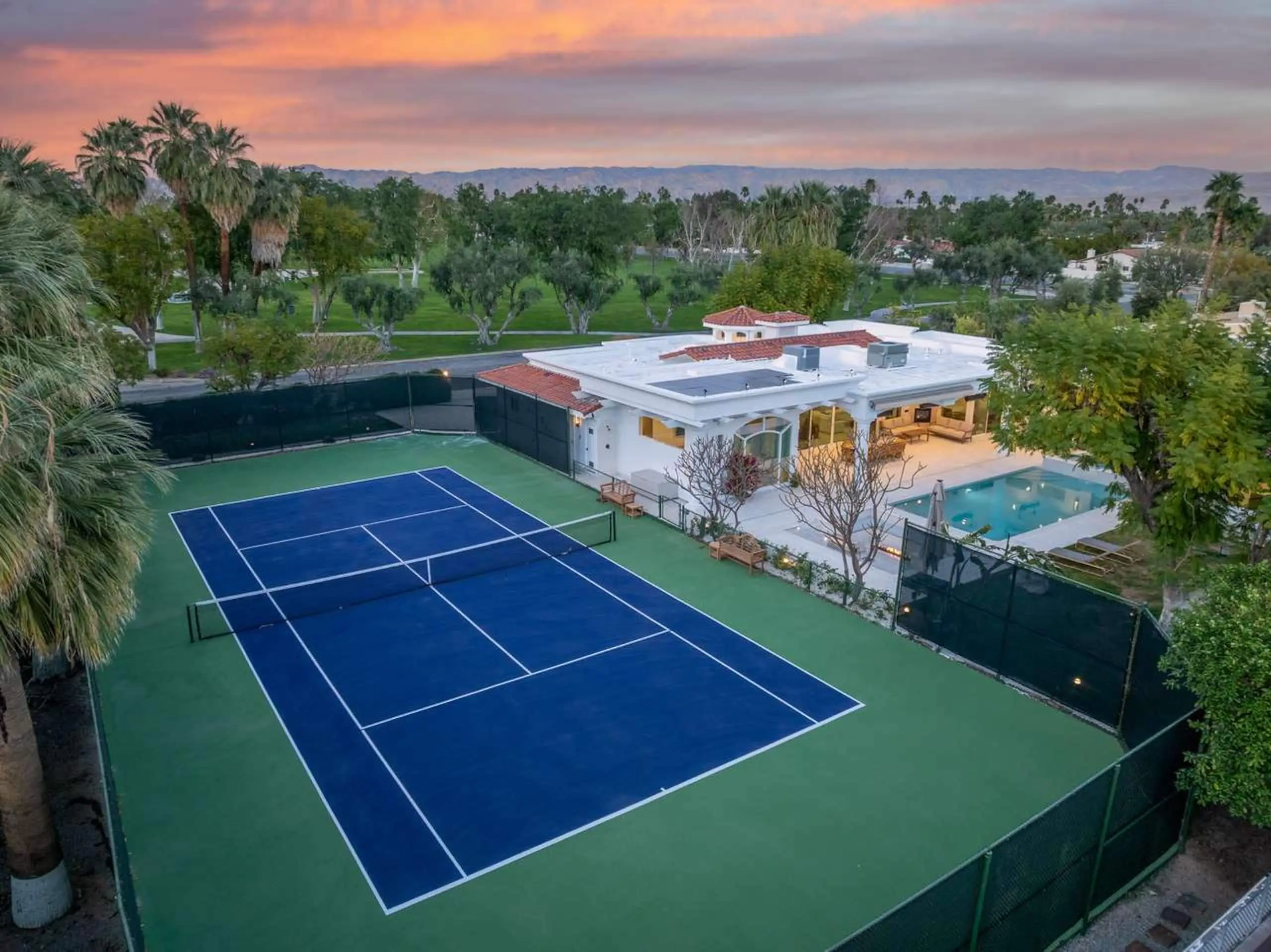 The whole property from the air: a full private tennis court, the house with its terracotta roof and colonnade, the pool, and the Coachella valley beyond.