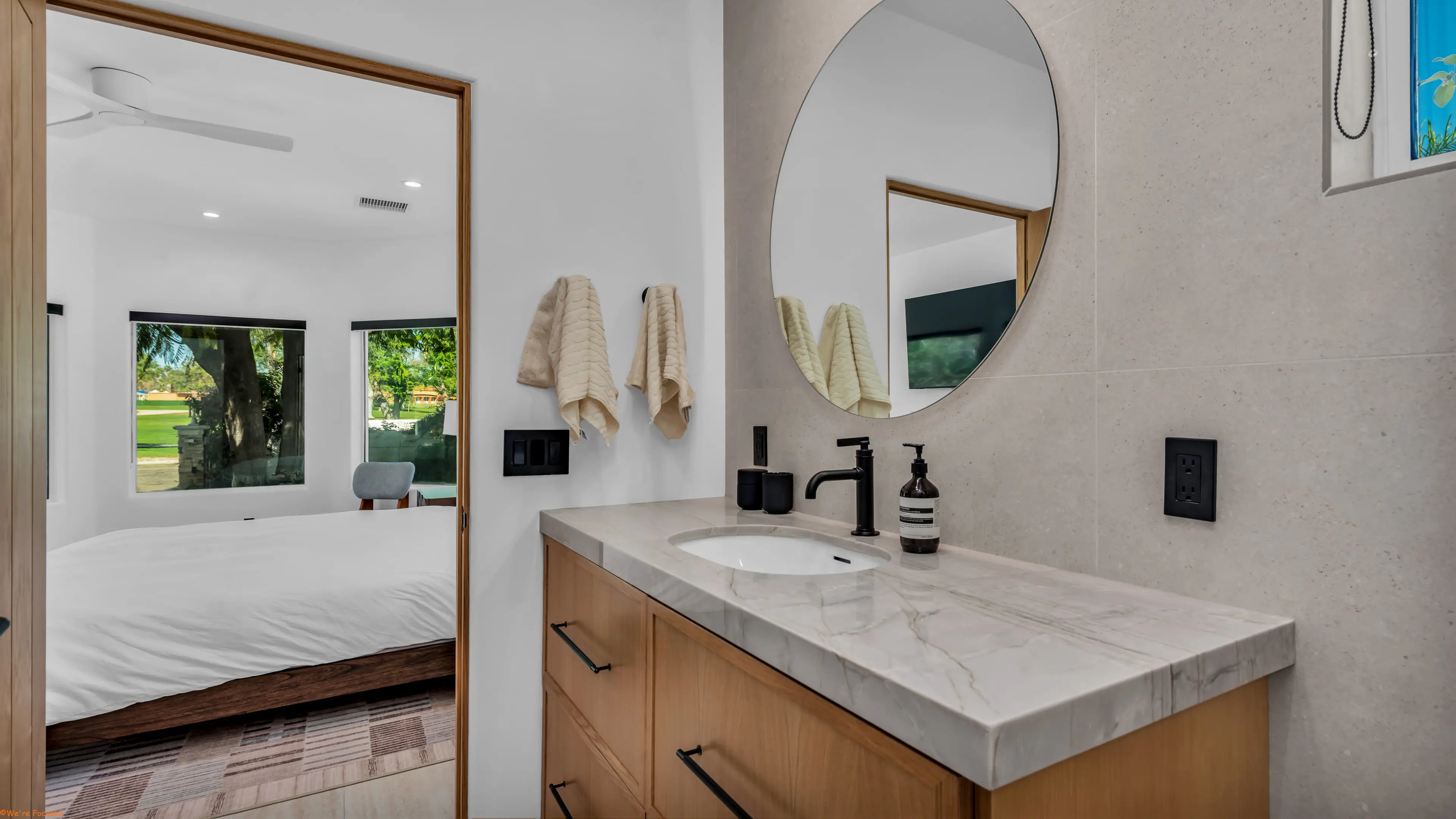 An oak vanity with a marble top, round mirror and a window through to the bedroom.