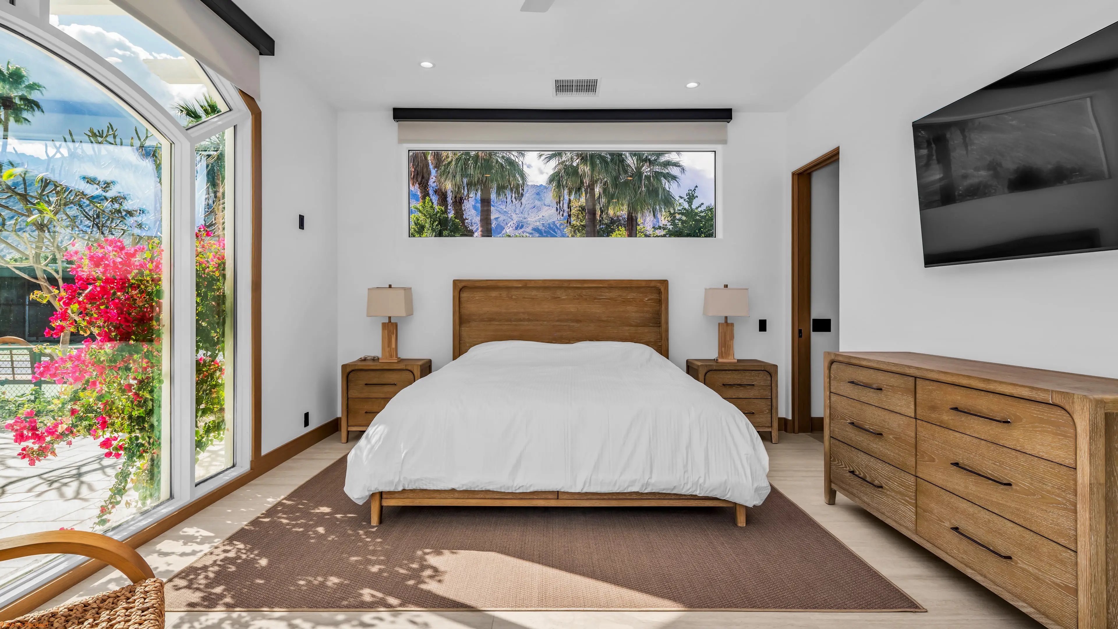 Bedroom with a corner window filled with bougainvillea and palms, and a long window over the bed.