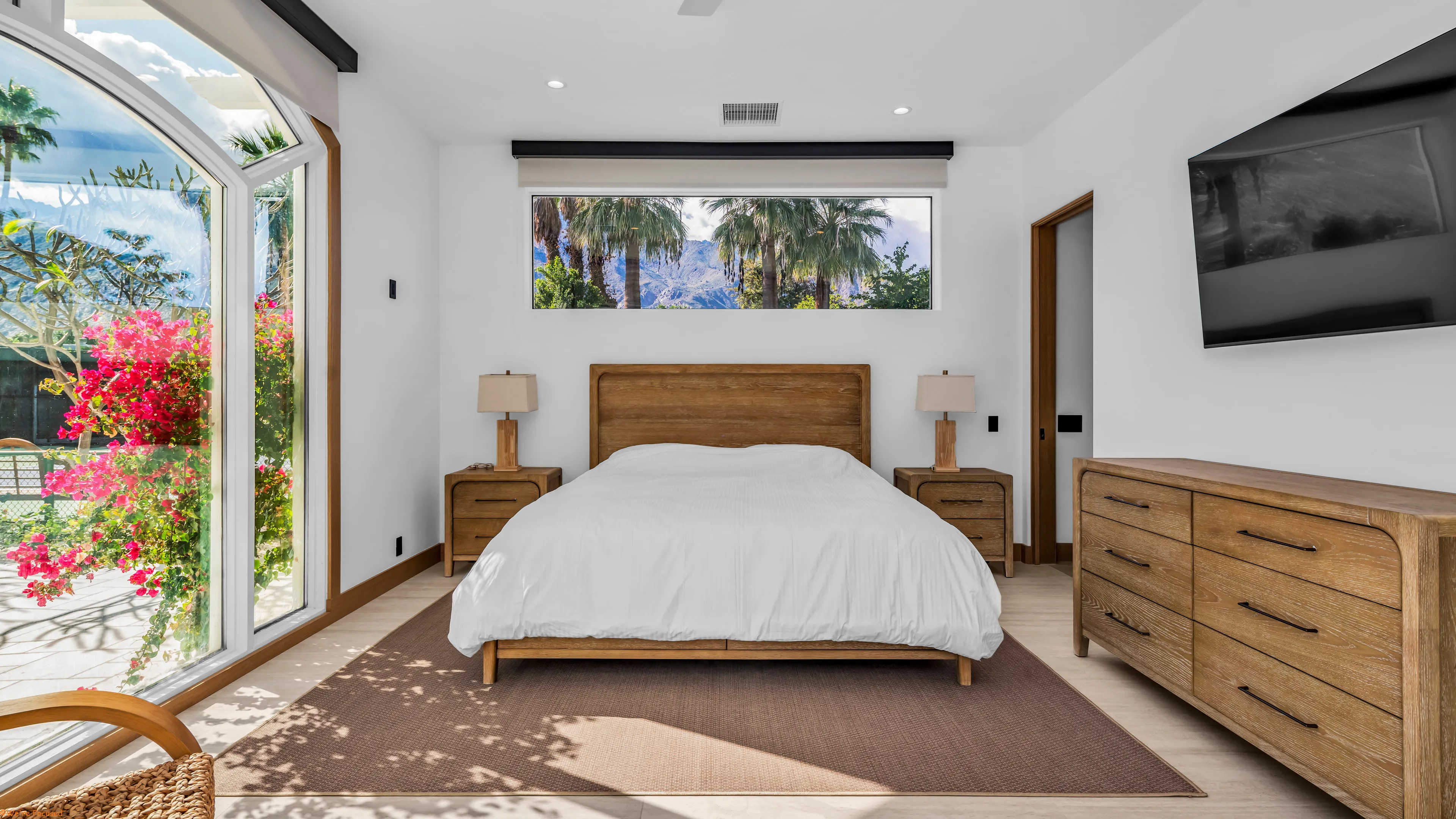 Bedroom with a corner window filled with bougainvillea and palms, and a long window over the bed.