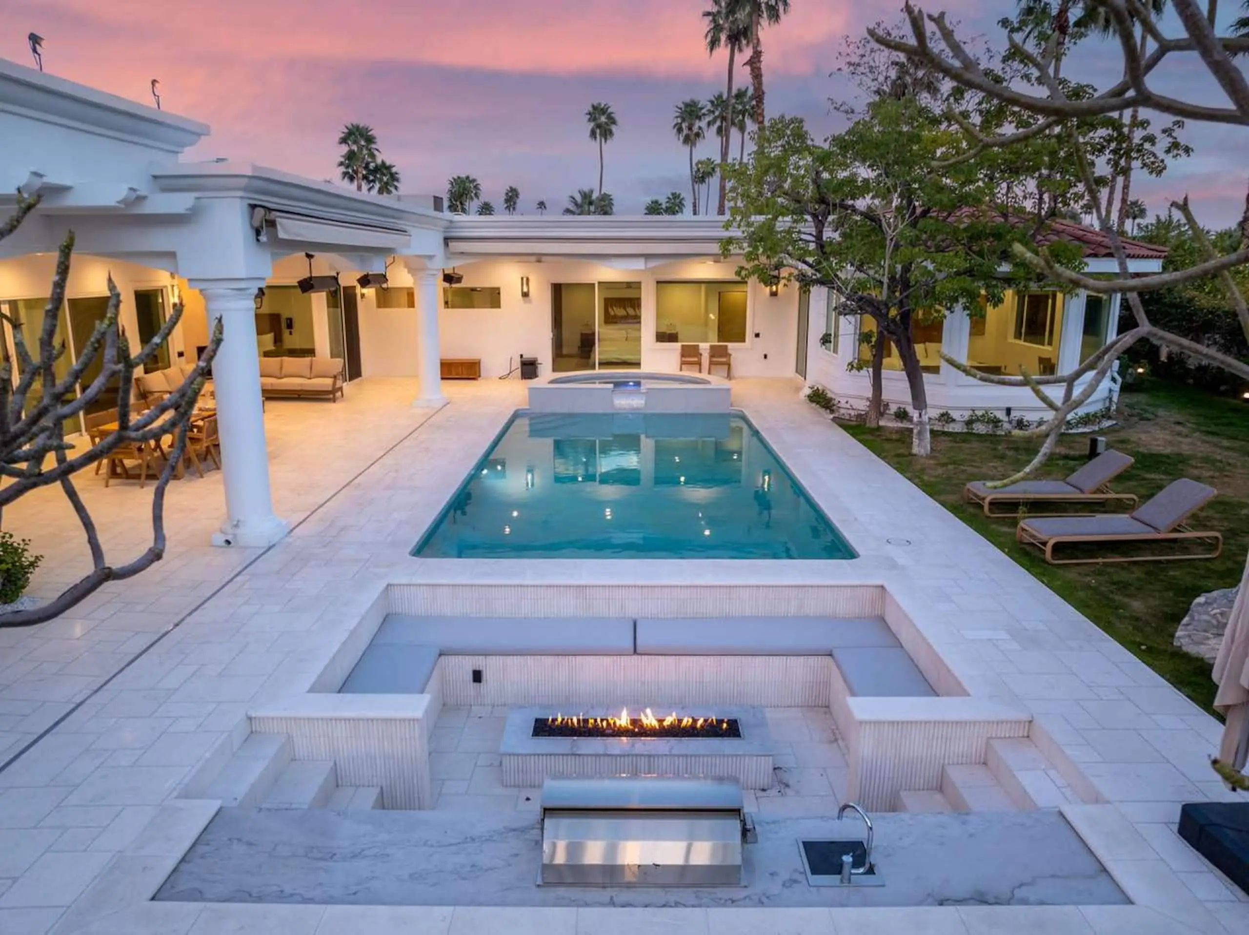 The courtyard at twilight: the lit pool, the spa, the sunken fire pit alight, and the colonnade against a pink sky.