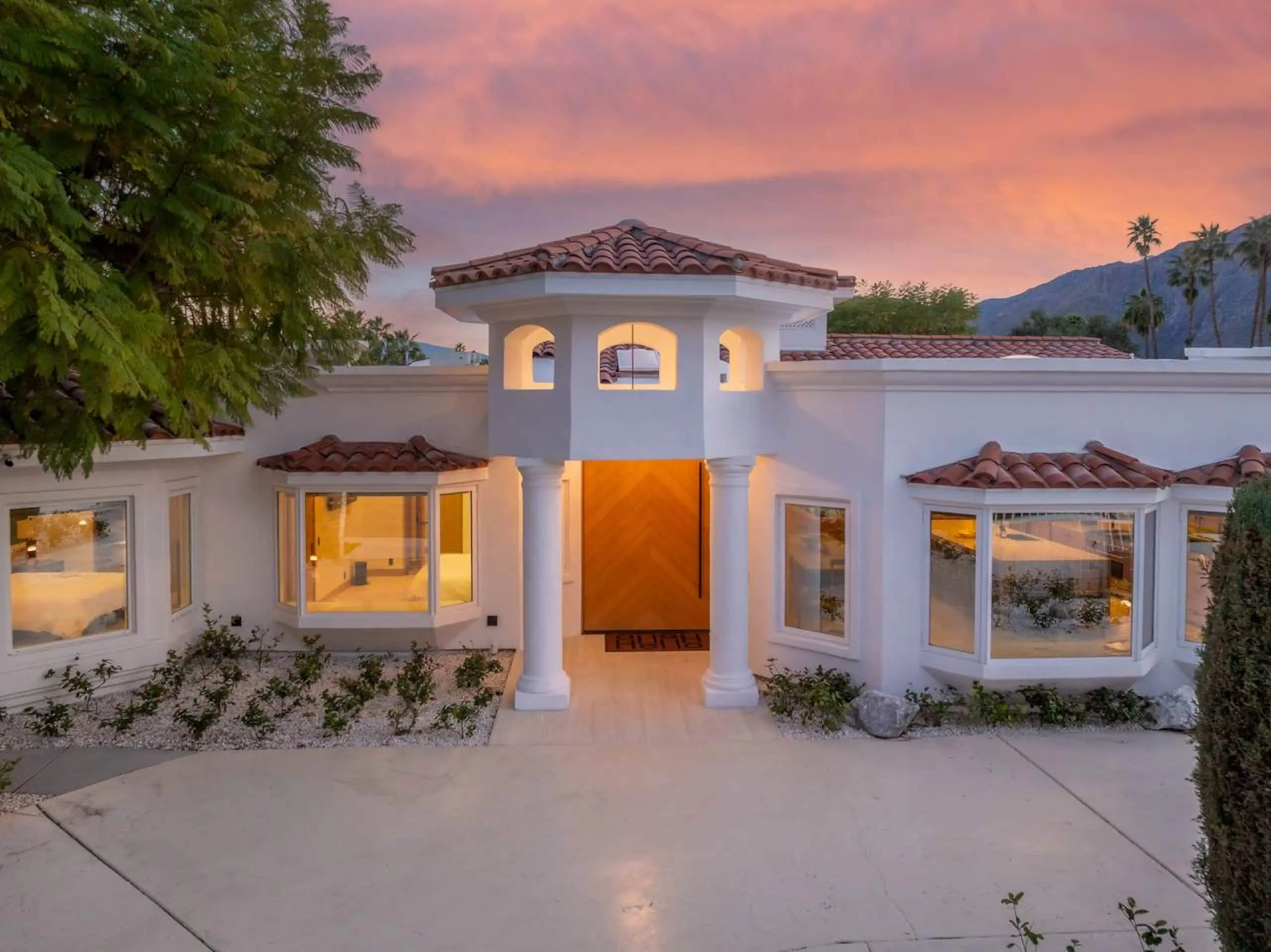 The front of the house at twilight, the chevron oak door lit from within between white columns, under a pink sky.