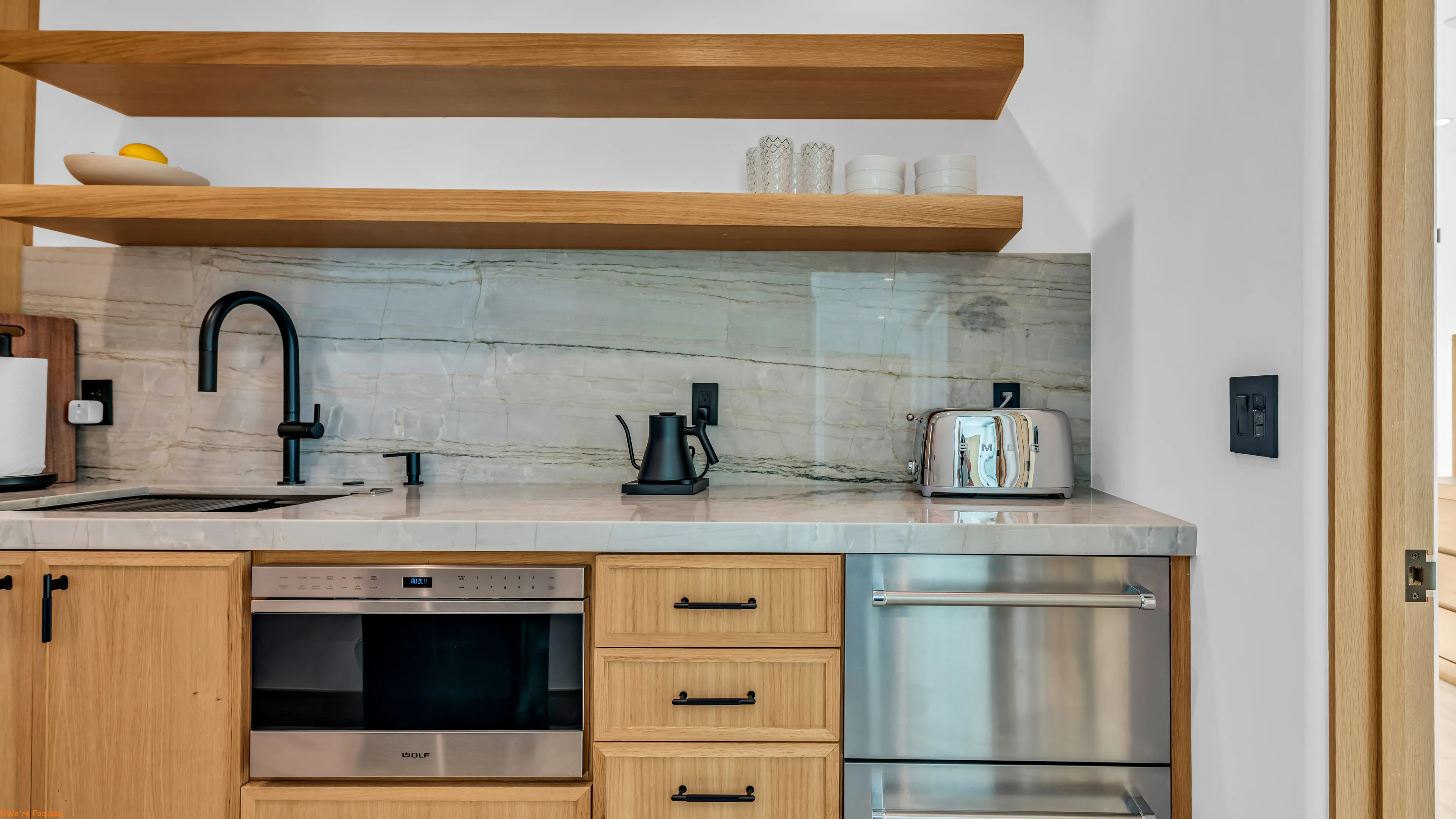 A counter run in pale veined stone under floating oak shelves, with cabinetry and built in units below.
